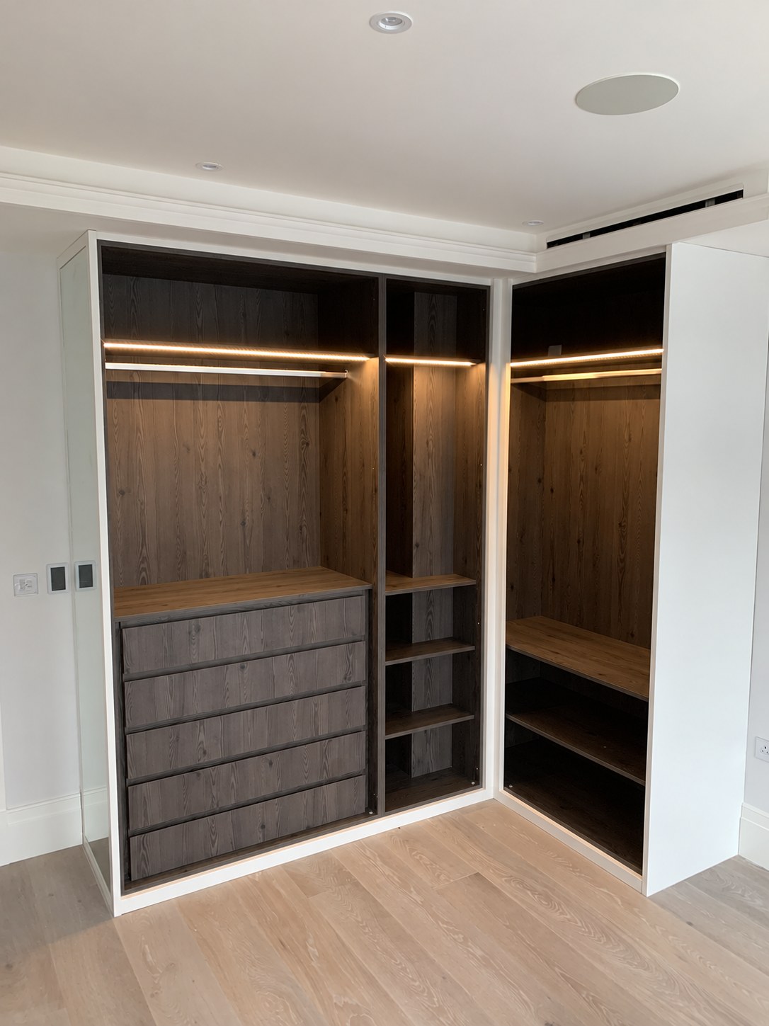 An L-shaped wardrobe with dark timber drawers, illuminated hanging space and a concealed AC outlet